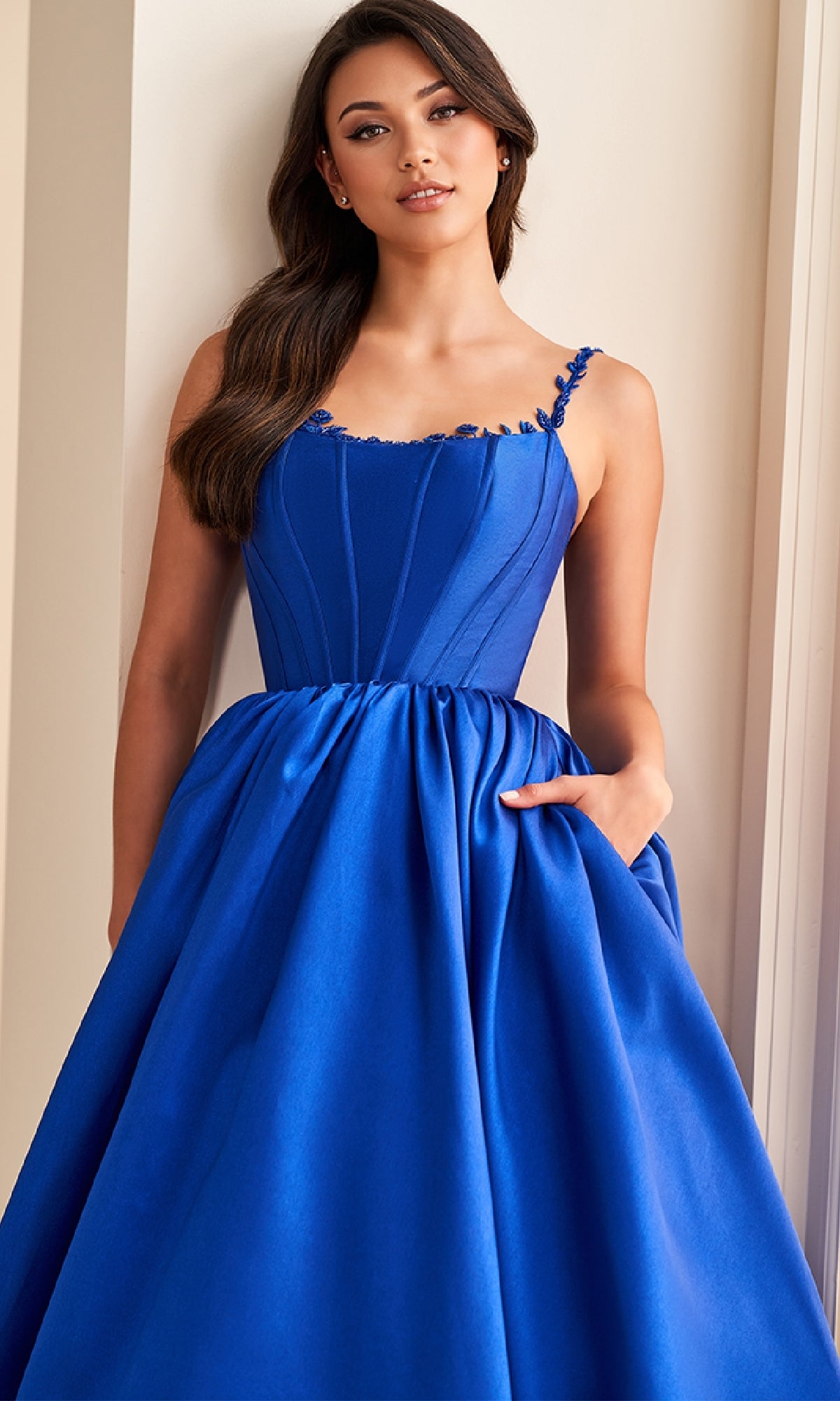 A woman stands indoors wearing the Ellie Wilde EW36221, a fitted royal blue long prom dress with a corset bodice, full A-line skirt, and thin straps, looking at the camera.